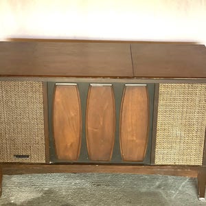 May include: A vintage wooden console stereo with a dark brown finish. The front features woven speaker covers and three vertical wooden panels. The stereo sits on four legs and has a hinged top. The overall design is mid-century modern.