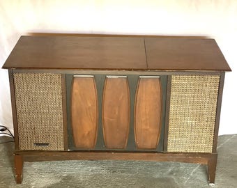 May include: A vintage wooden console stereo with a dark brown finish. The front features woven speaker covers and three vertical wooden panels. The stereo sits on four legs and has a hinged top. The overall design is mid-century modern.