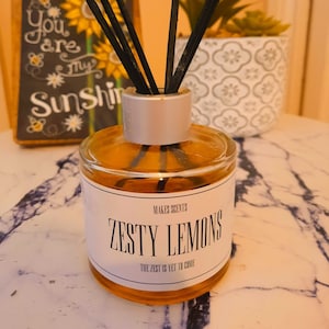 May include: A clear glass bottle with a silver lid and a white label that reads "Makes Scents Zesty Lemons The Zest is Yet to Come". The bottle is filled with a yellow liquid and has black diffuser sticks.