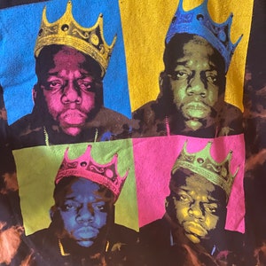 May include: A black t-shirt with a tie-dye pattern featuring four images of the rapper The Notorious B.I.G. wearing a crown. Each image is in a different color: blue, yellow, green, and pink.