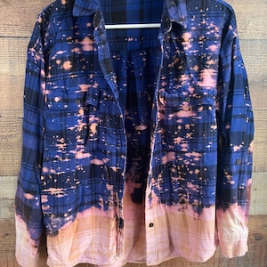 May include: A blue and black plaid flannel shirt with a bleach-splattered design. The shirt has a faded pink and white ombre effect at the bottom.