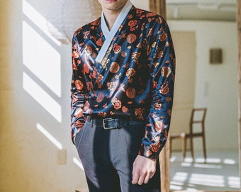 May include: A man wearing a black baseball cap, a blue and red patterned shirt with a white collar, black pants, and a black belt. The shirt has a traditional Korean design.