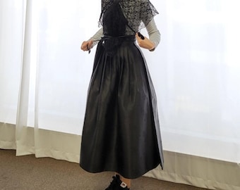 May include: A black skirt with a lace overlay and a black belt. The skirt is pleated and has a long, flowing silhouette. The model is wearing black sneakers.