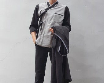 May include: A gray and black shirt with a long sleeve and a black sash tied across the front. The shirt has a pocket on the left side. The person is wearing black pants and holding a black jacket.