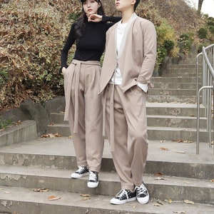 May include: A couple wearing matching brown pants and jackets pose on a set of stairs. The woman is wearing a black turtleneck and a white cap. The man is wearing a white shirt underneath his jacket.