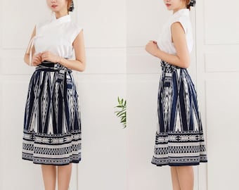 May include: A woman is wearing a white sleeveless top with a blue and white patterned skirt. The skirt has a geometric design and is knee-length. She is wearing black heeled sandals.