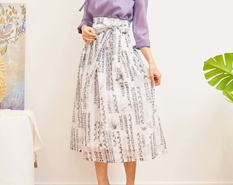 May include: A white skirt with a black and white print of Korean characters and flowers. The skirt is tied at the waist with a white sash. The skirt is worn with a light purple blouse and black heels.