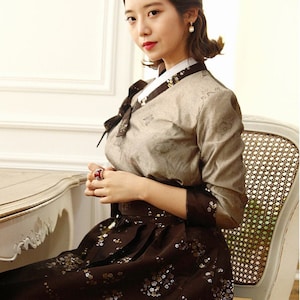 Może przedstawiać: Kobieta w tradycyjnej koreańskiej sukni hanbok. Hanbok to beżowa bluzka z kwiatowym wzorem i brązowa spódnica z kwiatowym wzorem w kolorze złotym i białym. Kobieta siedzi na krześle z plecionym oparciem.