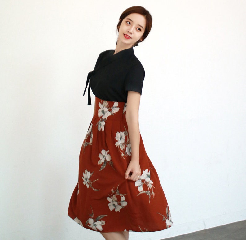 May include: A woman wearing a black short-sleeved top with a tied neckline and a rust-colored skirt with white floral print.