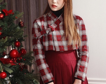 May include: A woman wearing a red and white plaid jacket with a red beret and a long red skirt. She is standing next to a Christmas tree.