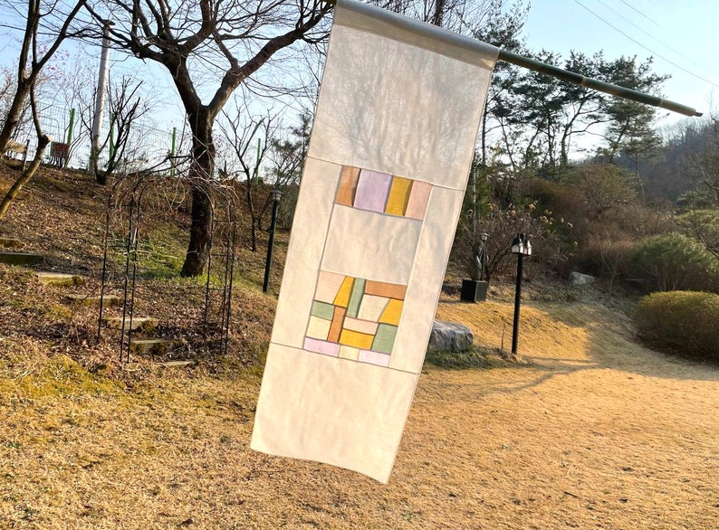 May include: A white fabric banner with a geometric design in shades of yellow, orange, green, and purple. The banner is hanging from a wooden pole.