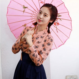 May include: A woman wearing a pink and floral patterned blouse and a dark blue skirt. She is holding a pink and white umbrella with floral designs.