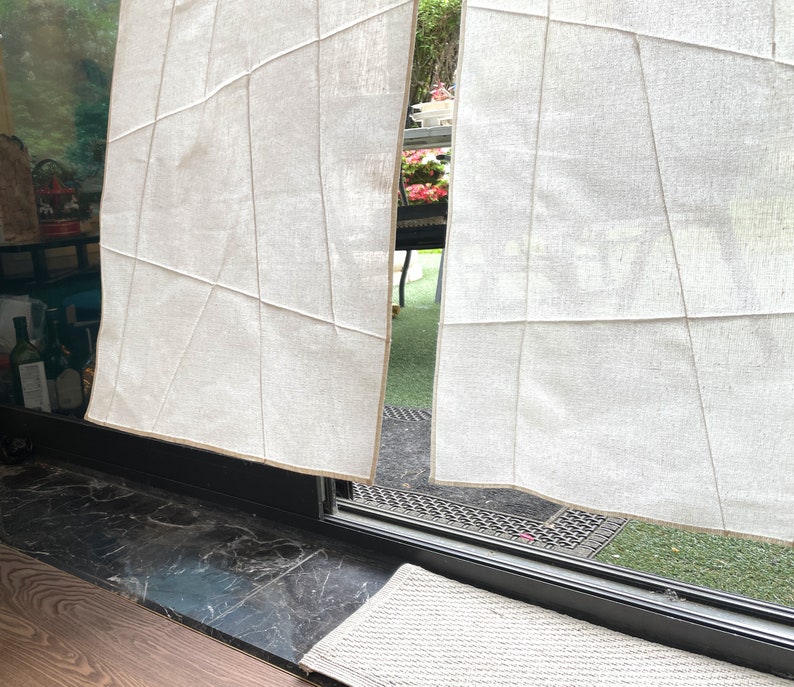 Op de afbeelding: Witte linnen gordijnen met een geometrisch patroon, hangend in een raam met uitzicht op een groen gazon.
