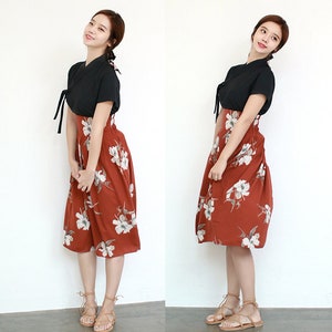 May include: A woman wearing a black short-sleeved top with a tied neckline and a rust-colored skirt with white floral print. She is wearing brown leather sandals with laces.