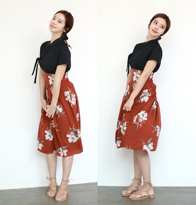 May include: A woman wearing a black short-sleeved top with a tied neckline and a rust-colored skirt with white floral print. She is wearing brown leather sandals with laces.