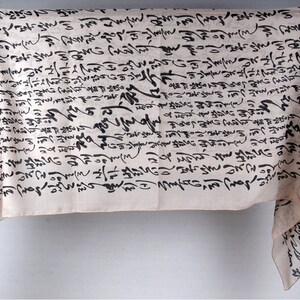 May include: A white fabric scarf with black calligraphy-style writing all over it. The scarf is draped over a stand.