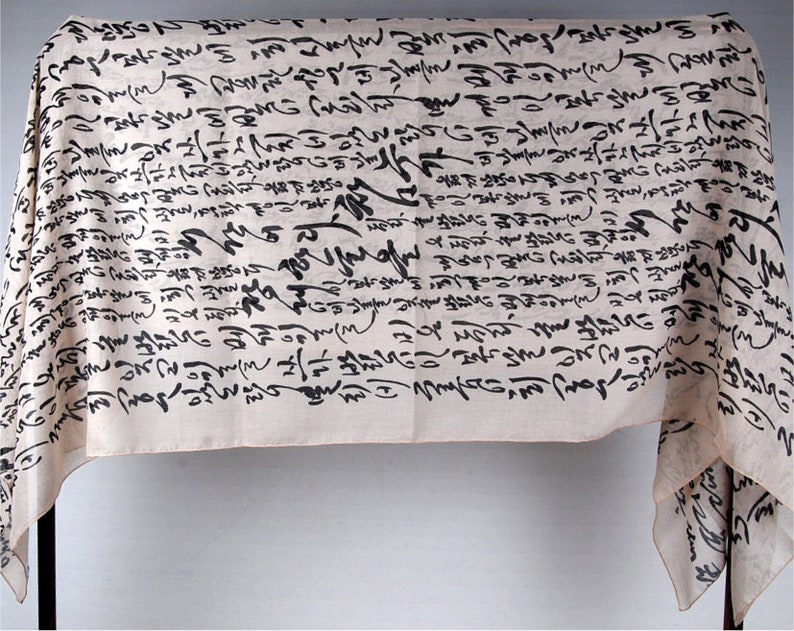 May include: A white fabric scarf with black calligraphy-style writing all over it. The scarf is draped over a stand.