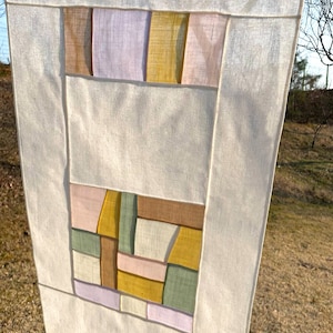 May include: A white fabric wall hanging with a geometric design. The design is made up of squares and rectangles in shades of pink, brown, yellow, and green.
