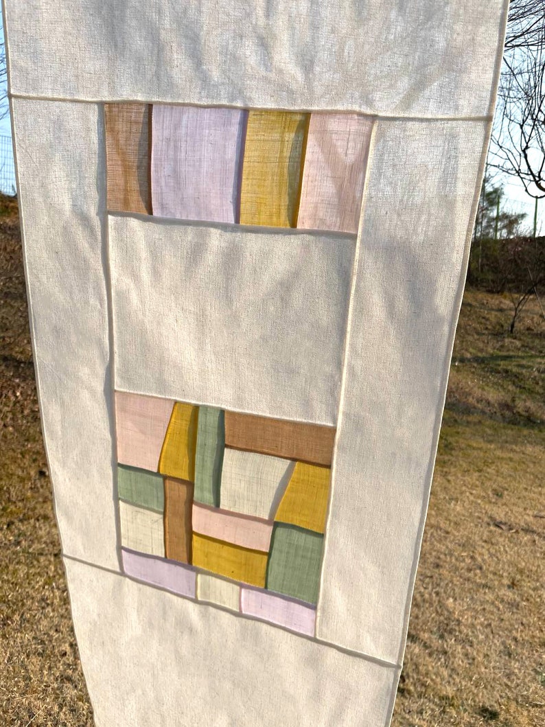 May include: A white fabric wall hanging with a geometric design. The design is made up of squares and rectangles in shades of pink, brown, yellow, and green.