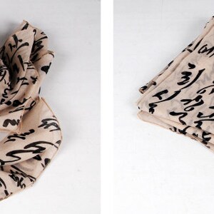 May include: A beige scarf with black calligraphy-style writing. The scarf is shown folded and laid flat.