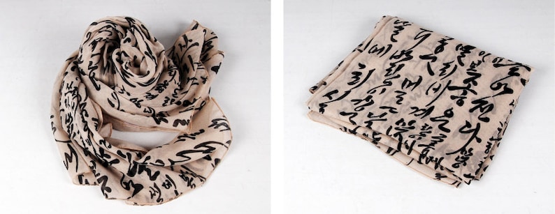 May include: A beige scarf with black calligraphy-style writing. The scarf is shown folded and laid flat.