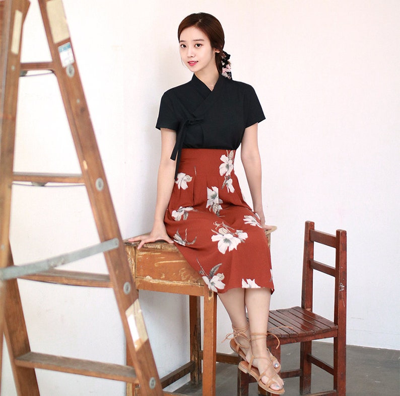 May include: A woman wearing a black short-sleeved top with a high neckline and a red floral skirt. She is sitting on a wooden table with a wooden chair beside her.