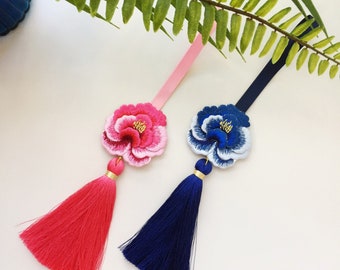 Könnte beinhalten: Zwei gestickte Blumenanhänger mit Quasten. Ein Anhänger ist rosa mit einer roten Quaste, der andere ist blau mit einer dunkelblauen Quaste.