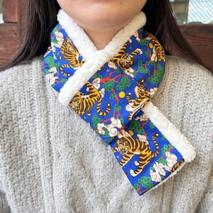 May include: A blue scarf featuring a repeating pattern of tigers, green pine trees, and white clouds. The scarf is lined with a thick, white, textured fleece material that creates a soft border along the edges of the blue fabric.