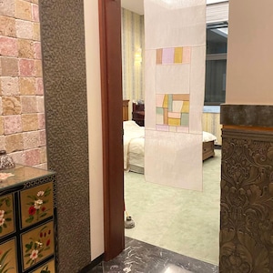 May include: A view of a room with a decorative screen made of white fabric with colorful geometric patterns. The screen is hanging in a doorway leading to another room. The room has a stone wall with a small decorative cabinet with floral designs.