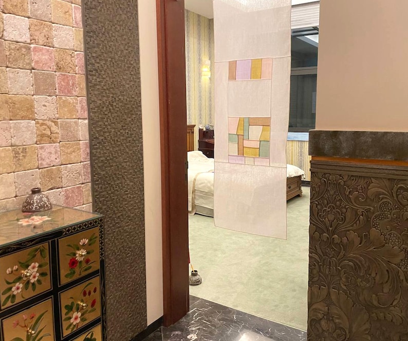 May include: A view of a room with a decorative screen made of white fabric with colorful geometric patterns. The screen is hanging in a doorway leading to another room. The room has a stone wall with a small decorative cabinet with floral designs.