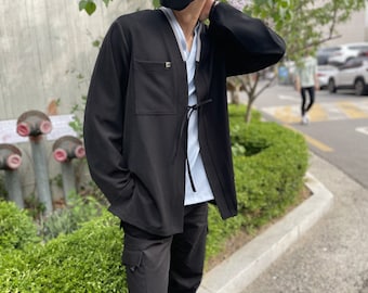 May include: Black jacket with a front pocket and a tie closure, worn over a light blue shirt. The jacket has long sleeves and a small gold-colored emblem on the pocket. The person is also wearing black cargo pants.