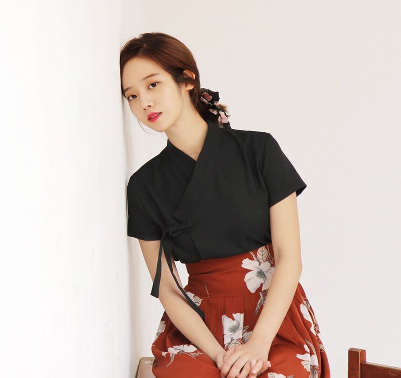 May include: A woman wearing a black short-sleeved top with a tied sash and a red floral skirt.