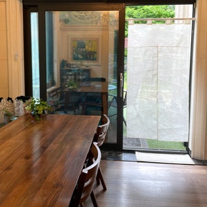 Op de afbeelding: Een houten eettafel met vier stoelen in een kamer met een schuifdeur van glas en een wit transparant gordijn. De tafel heeft een natuurlijke houten afwerking en de stoelen zijn bruin.