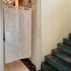 Op de afbeelding: Een witte linnen roomdivider met een geometrisch patroon hangt aan een witte stang. De divider bevindt zich in een deuropening die leidt naar een trap met donkere marmeren treden.