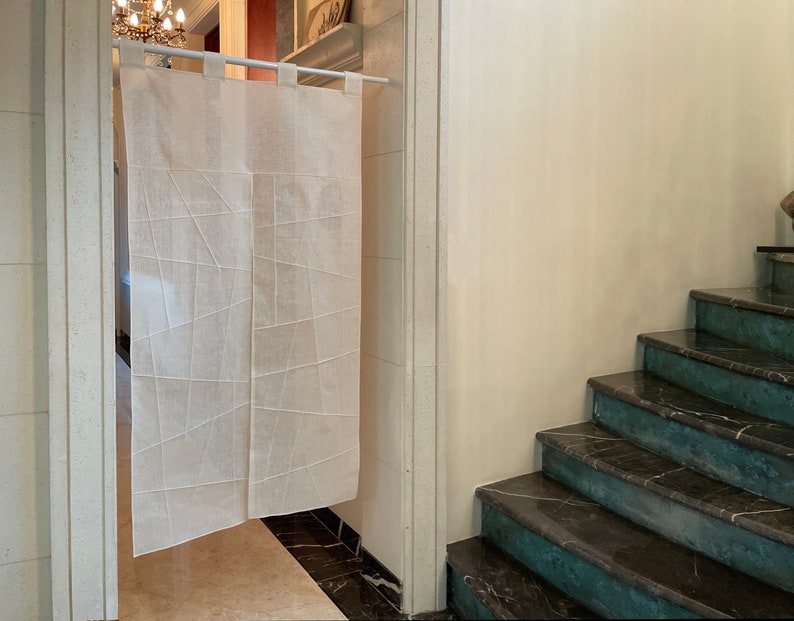Op de afbeelding: Een witte linnen roomdivider met een geometrisch patroon hangt aan een witte stang. De divider bevindt zich in een deuropening die leidt naar een trap met donkere marmeren treden.