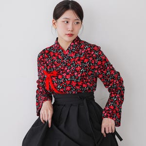 Może przedstawiać: Tradycyjny koreański strój hanbok. Góra to bluzka z długim rękawem z żywym czerwonym kwiatowym wzorem na czarnym tle, z czerwoną wstążką. Spódnica to pełna, zwiewna czarna spódnica.