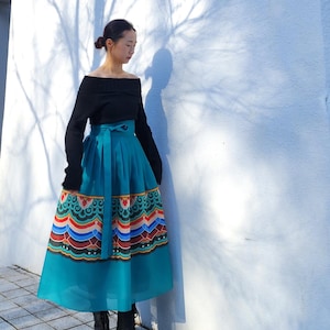 Puede incluir: Un top negro de hombros descubiertos y manga larga combinado con una falda midi de color verde azulado de talle alto. La falda presenta una cintura ancha con cinturón de lazo y un patrón gráfico colorido de varias capas a lo largo del dobladillo.