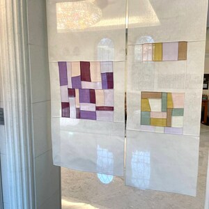 May include: Two white panels with geometric patterns in shades of purple, pink, brown, yellow, and green. The panels are made of fabric and are hanging on a wall.