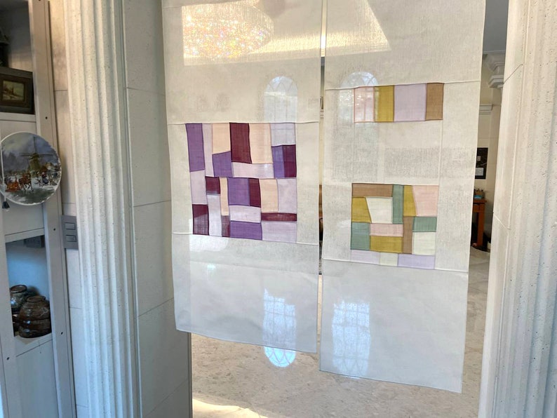 May include: Two white panels with geometric patterns in shades of purple, pink, brown, yellow, and green. The panels are made of fabric and are hanging on a wall.