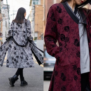 Puede incluir: Un abrigo largo con un estampado floral en negro y gris, con un cinturón negro y un cuello ancho. El abrigo se lleva sobre pantalones y botas negras. También se muestra un segundo abrigo en burdeos con un estampado floral negro.