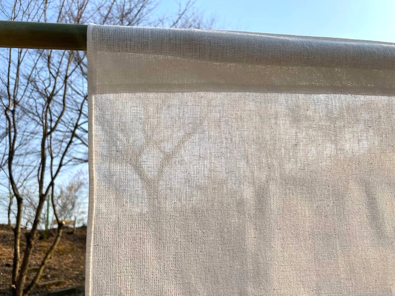 May include: A white cotton fabric hanging on a wooden rod. The fabric is thin and translucent, allowing light to pass through it.