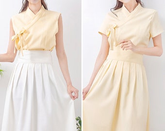 May include: Two women wearing a yellow top with a white skirt. The top is a traditional Korean style with a V-neck and a tie at the front. The skirt is a simple A-line style with pleats.