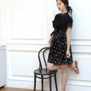 May include: A woman wearing a black dress with a floral print skirt and brown sandals. She is standing next to a black chair with her leg resting on the seat.