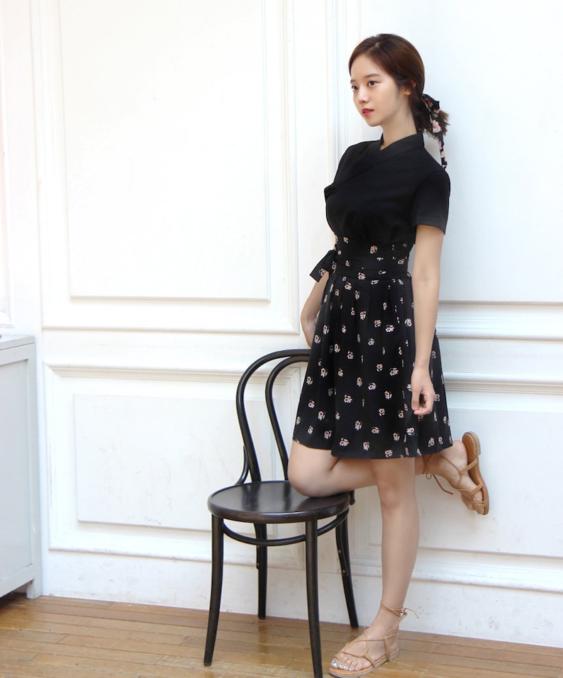 May include: A woman wearing a black dress with a floral print skirt and brown sandals. She is standing next to a black chair with her leg resting on the seat.