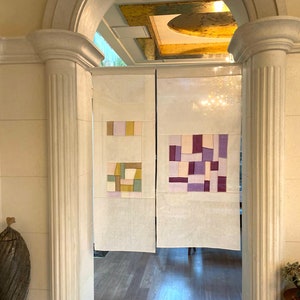 May include: A pair of white fabric room dividers with geometric patterns in shades of purple, pink, and yellow. The dividers are hanging in a doorway with a white trim.