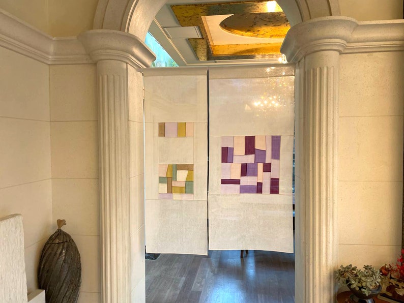 May include: A pair of white fabric room dividers with geometric patterns in shades of purple, pink, and yellow. The dividers are hanging in a doorway with a white trim.