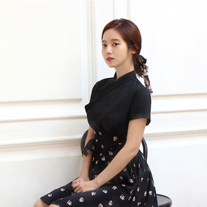 May include: A woman wearing a black dress with a floral print skirt. The dress has a short sleeve and a high neckline. She is sitting on a black chair in front of a white wall.