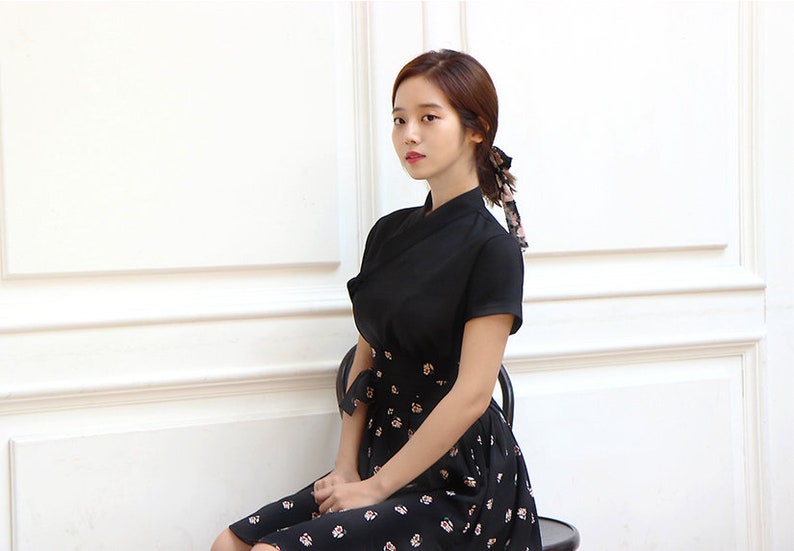 May include: A woman wearing a black dress with a floral print skirt. The dress has a short sleeve and a high neckline. She is sitting on a black chair in front of a white wall.