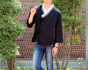 May include: A black and white traditional Korean shirt with long sleeves and a tied sash. The shirt is worn over blue jeans.