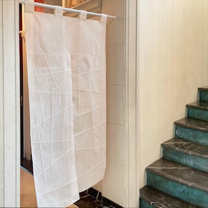 Op de afbeelding: Een wit linnen gordijn met een geometrisch patroon hangt in een deuropening. Het gordijn bestaat uit twee panelen, elk met een ander patroon. Het gordijn hangt aan een witte stang.